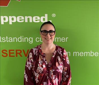 Female employee Sarah Raymond standing in front of a green wall with the SERVPRO mission statement